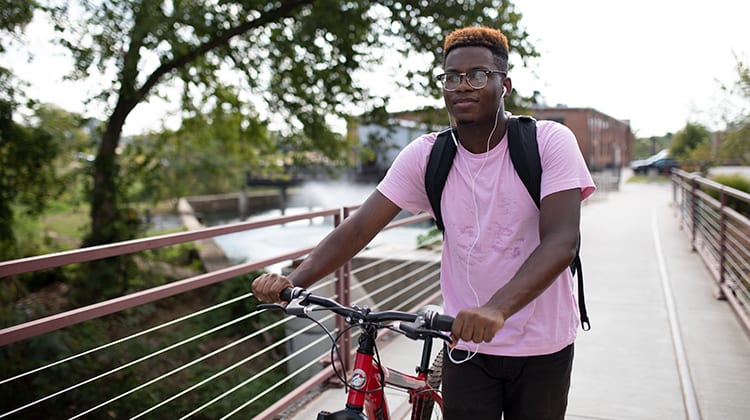 A young man walks on a college campus with his bike.