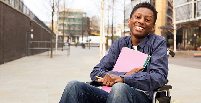 A student who uses a wheelchair holds folders in his arms and smiles.