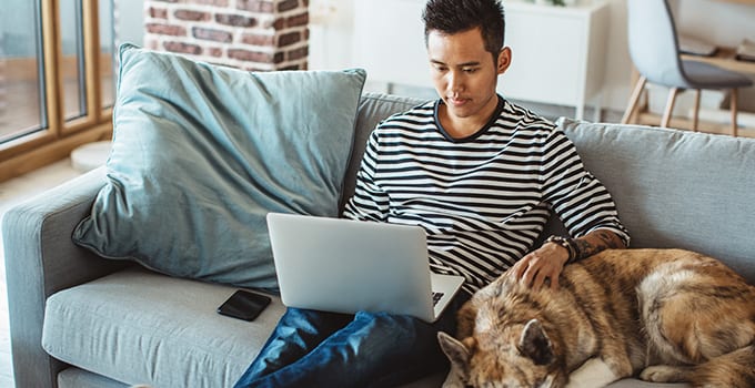 A student sits on a couch with a laptop on his lap and a dog laying next to him.